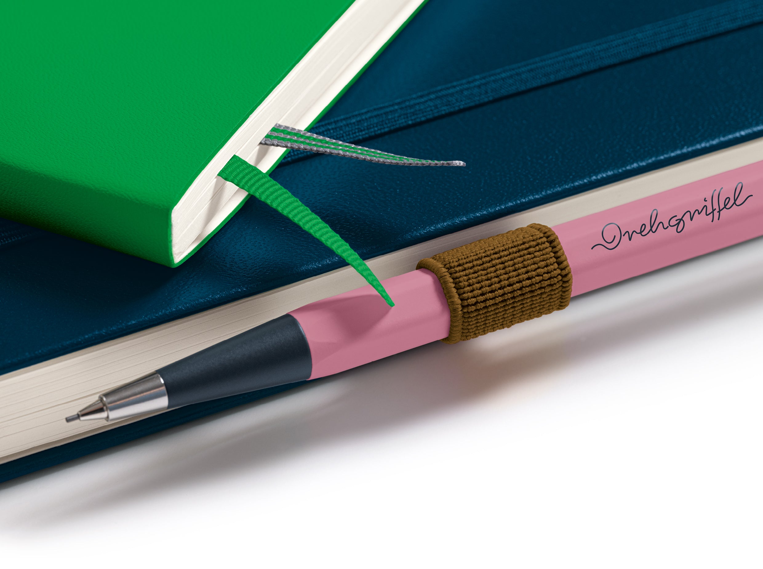 A pink pen with a brown elastic loop and black script logo, resting on a dark blue notebook next to a green one, on a white background.