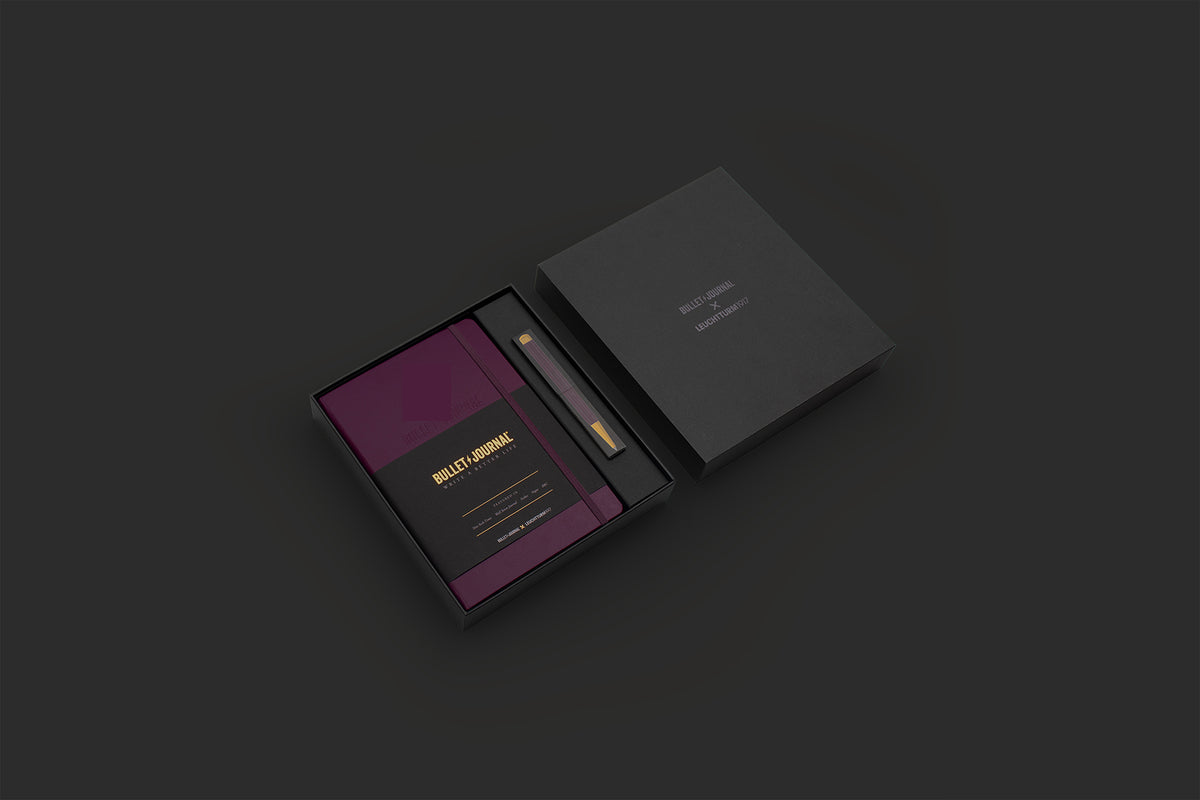 A premium black presentation box shown open on a dark charcoal background, revealing a deep purple insert card with gold text and a slim gold-and-black accessory inside.
