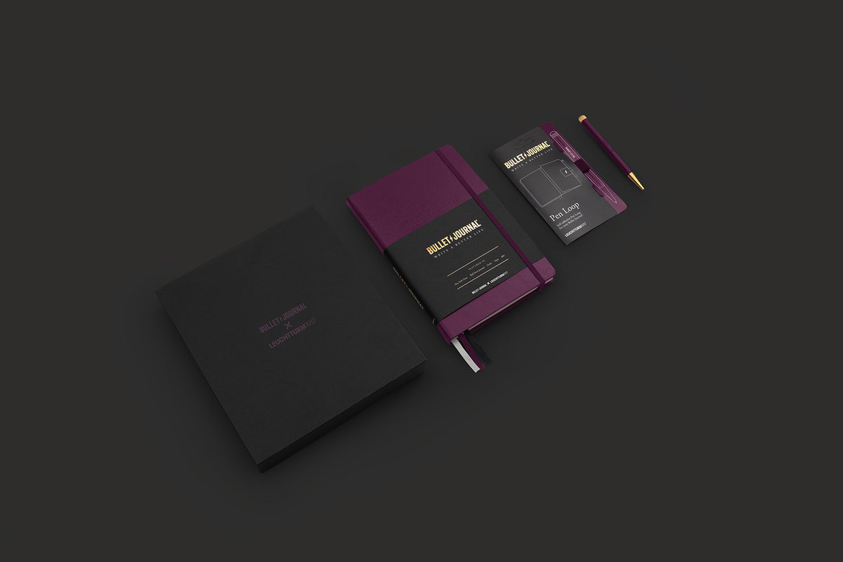A sleek stationery set on a dark charcoal background, featuring a black hardcover notebook, a purple and black slim notebook with gold lettering, a matching black card, and a purple pencil.
