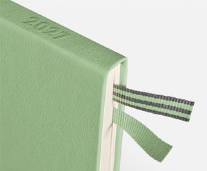 A close-up of a sage green textured 2027 journal or planner, showing the embossed year on the cover and a matching green and gray striped ribbon bookmark.