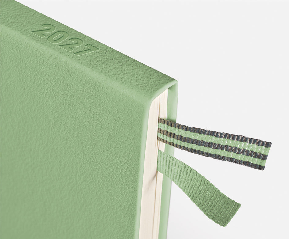A close-up of a sage green textured 2027 journal or planner, showing the embossed year on the cover and a matching green and gray striped ribbon bookmark.