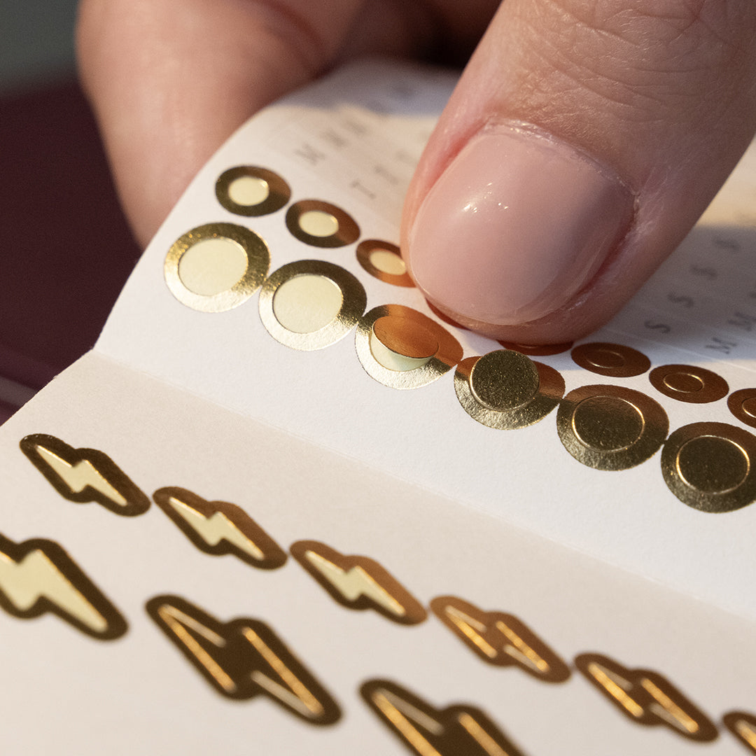 A close-up of a hand pressing down shiny gold oval stickers onto a white sheet, with more gold sticker shapes visible below.