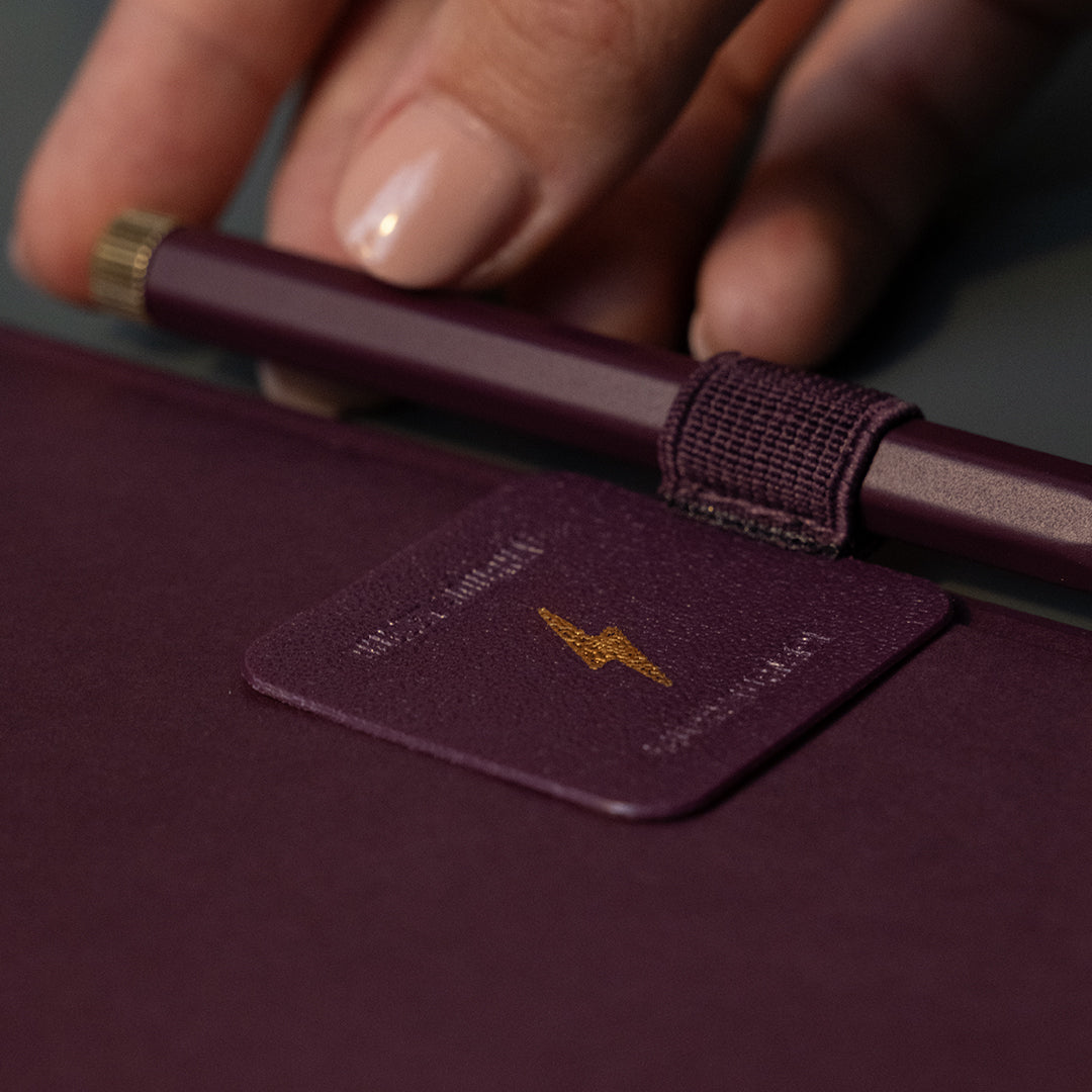 A close-up of a hand holding a dark plum pen over a matching dark plum folio or notebook cover with a small square leather tag featuring a gold lightning bolt logo.