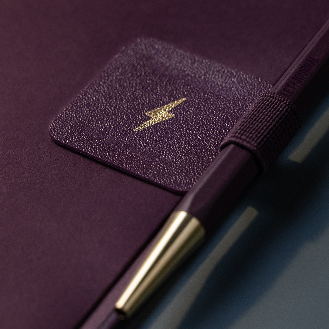 A close-up of a dark purple textured notebook or journal with a matching elastic closure band and a gold lightning bolt logo on a square patch, alongside a matching dark purple pen with a gold accent.
