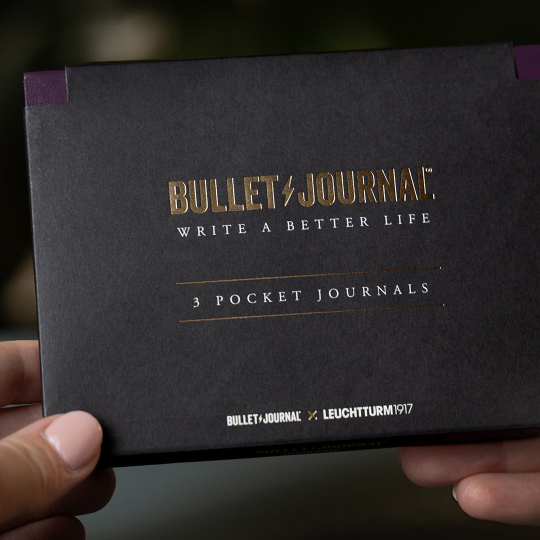A person's hands holding a black Bullet Journal 3 Pocket Journals box with gold foil lettering, set against a blurred dark background.