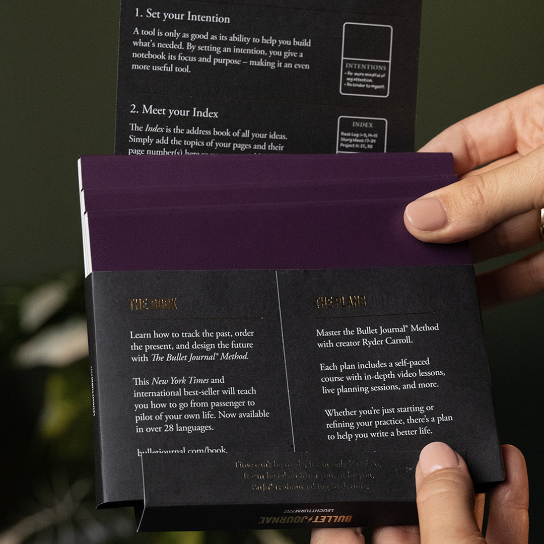 A person's hands holding open a dark purple and black Bullet Journal set, showing the inner pages with gold foil text and a dark olive green background.