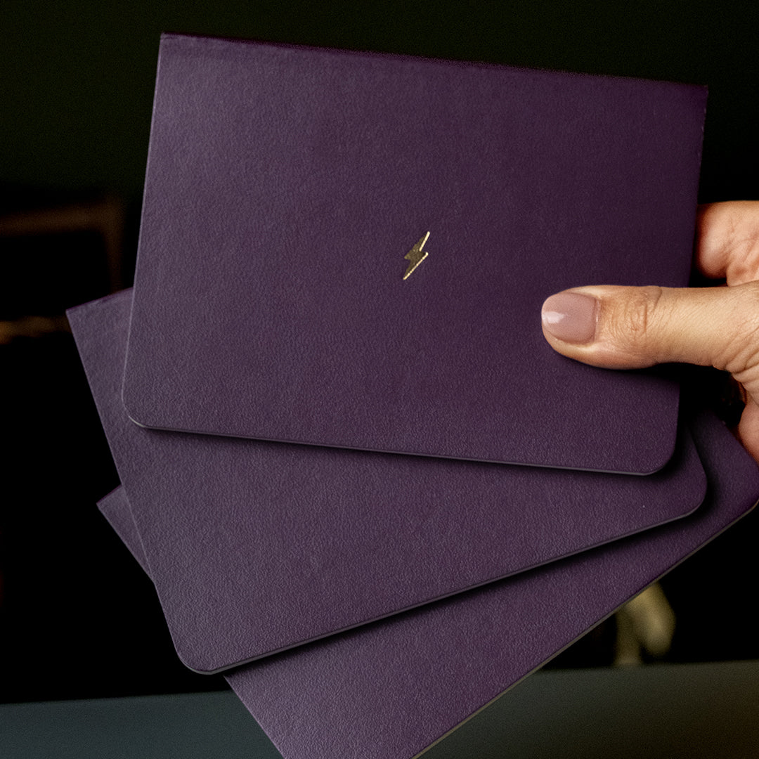A hand holding three stacked dark purple textured notebooks or card sleeves with a small gold lightning bolt logo on the top one, against a dark background.