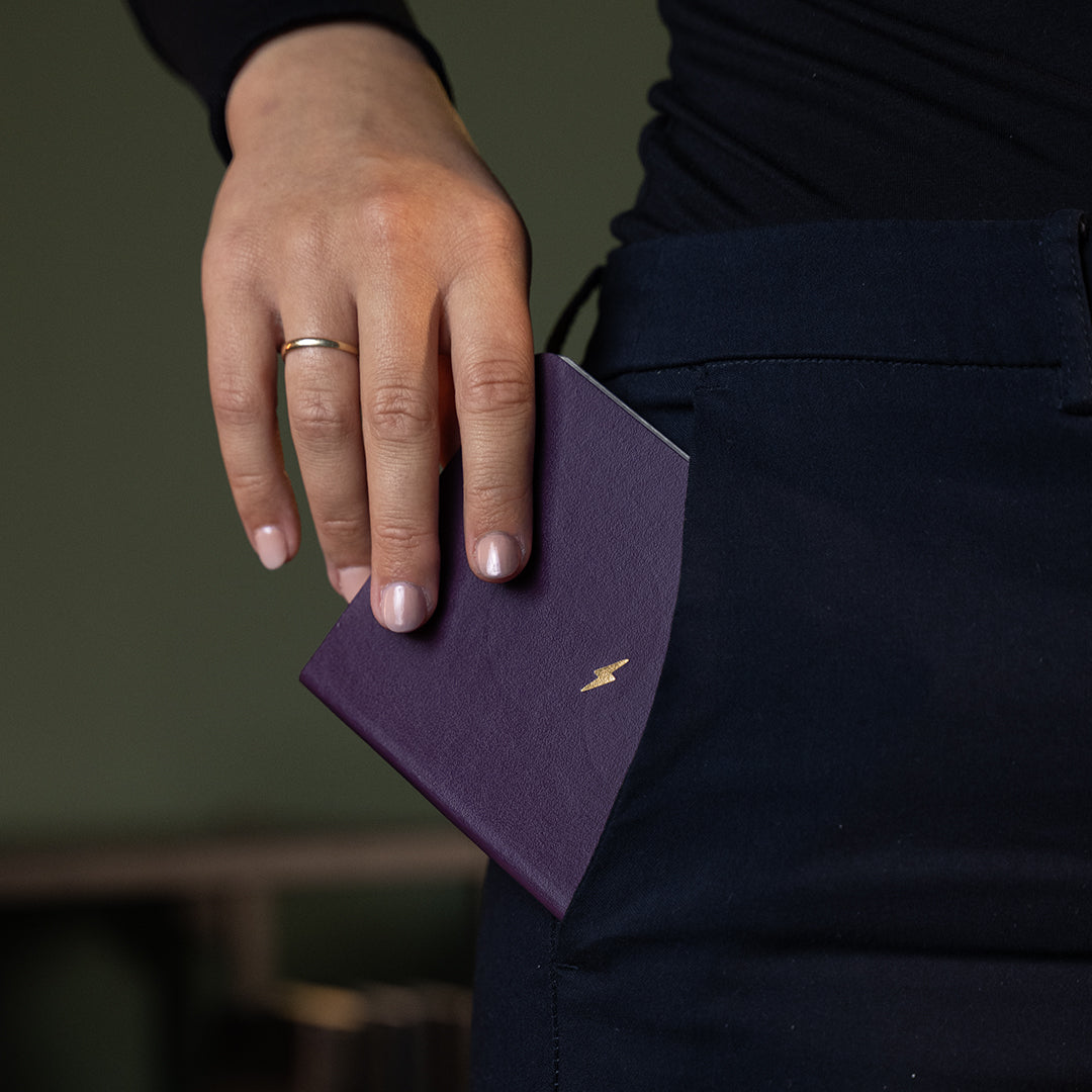 A close-up of a person's hand holding a small purple notebook or passport cover with a gold lightning-bolt logo, tucked into the back pocket of dark navy trousers.