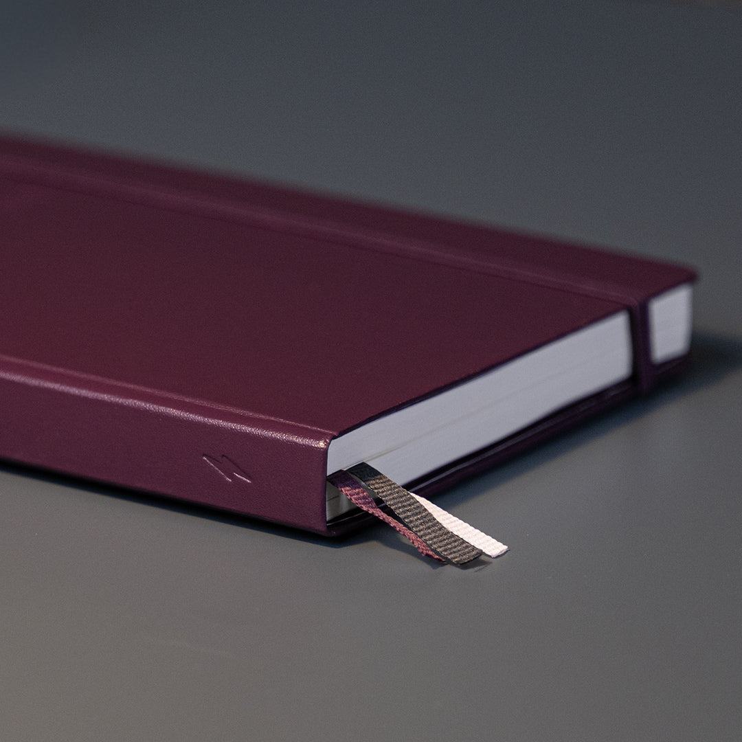 A close-up of a dark burgundy hardcover notebook or journal lying on a smooth gray surface, with a striped paper bookmark or page divider peeking out from the edge.