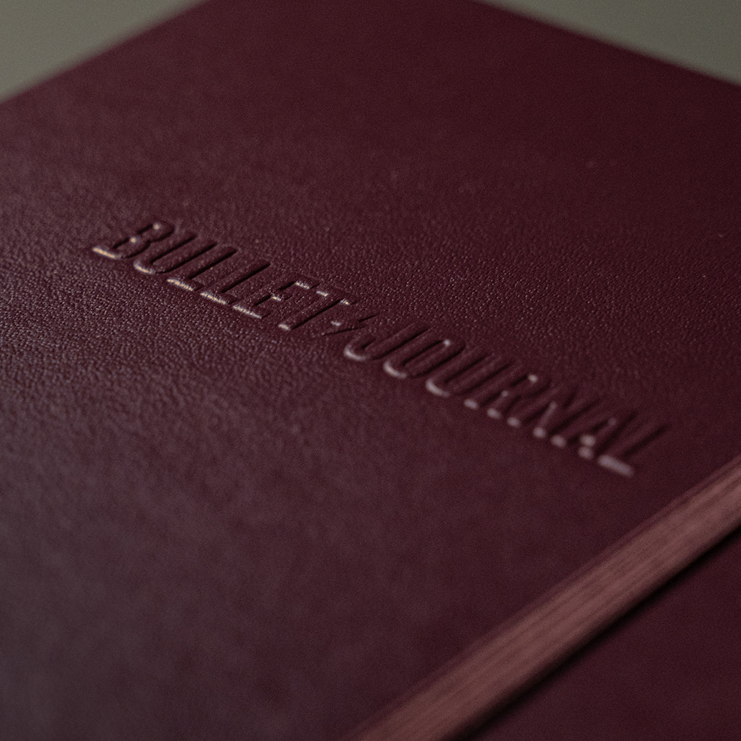 A close-up of a burgundy leather journal cover with the words 'BULLET JOURNAL' embossed on the front.