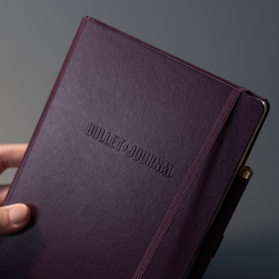 A close-up of a hand holding a dark purple Bullet Journal notebook with an embossed logo and an elastic closure band, set against a dark gray background.