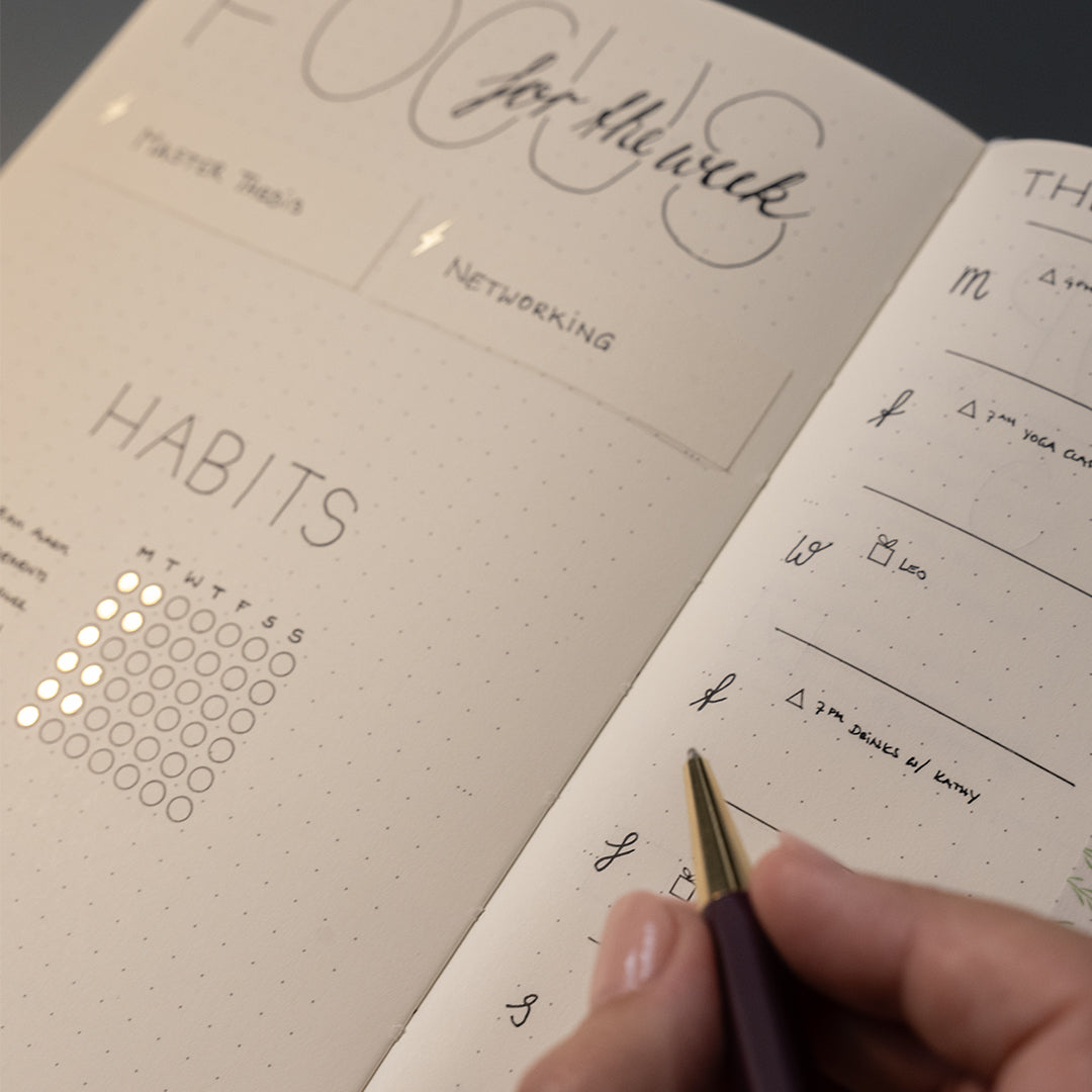 An open bullet journal with a hand holding a pen, showing pages with handwritten notes, habit tracking, and decorative script text on a dark background.