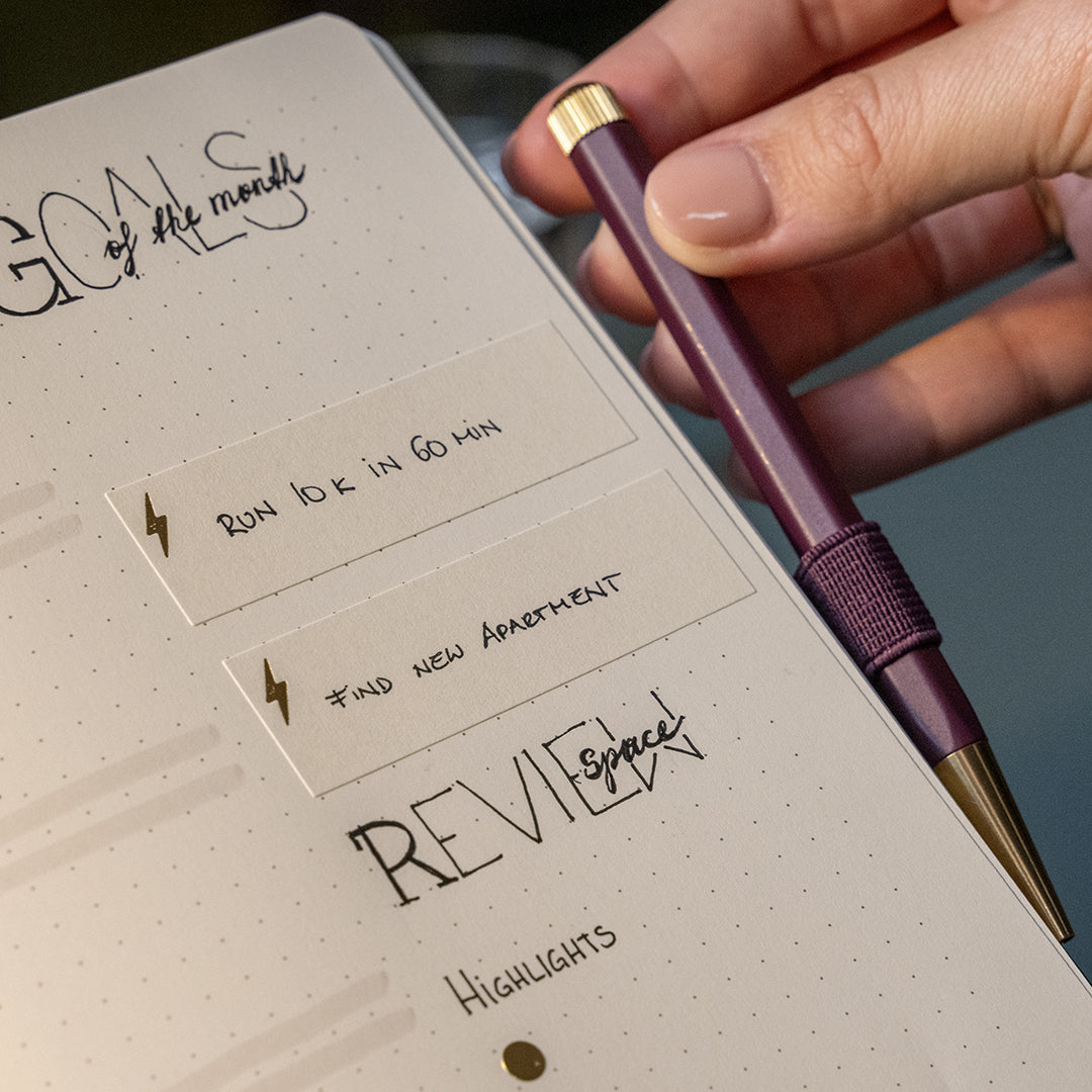 A close-up of a hand holding a metallic purple pen with gold accents next to an open planner or notebook with handwritten goals and review sections.