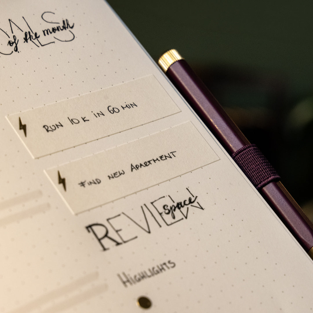 A close-up of a planner or notebook page with handwritten notes, sticky notes, and a metallic purple pen with a gold cap resting diagonally across the page.