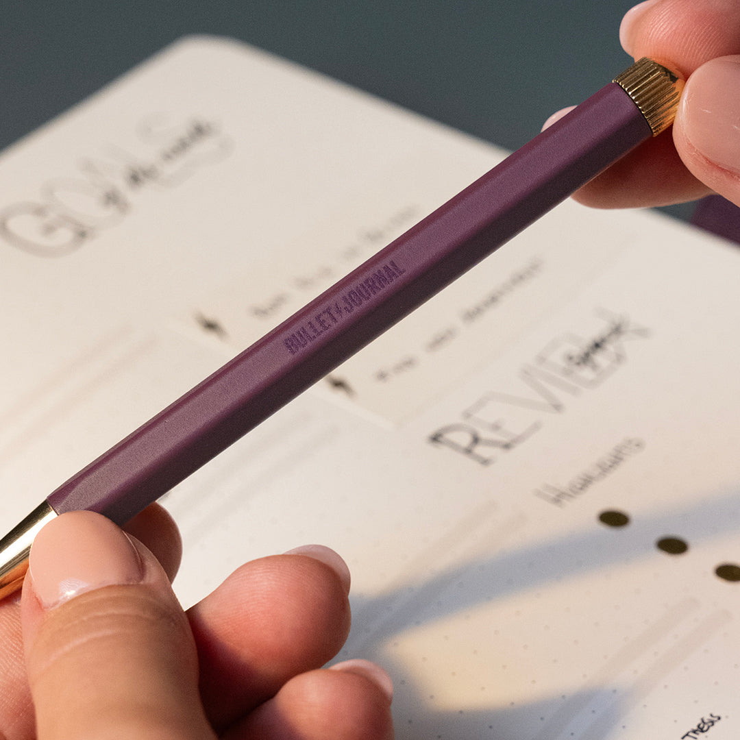 A close-up of a hand holding a purple bullet journal pen with gold accents over an open planner or notebook.
