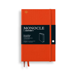 An orange Monocle accordion notebook by Leuchtturm1917, featuring a black paper band around the middle with white text and a small white circular button on the side, shown on a white background.