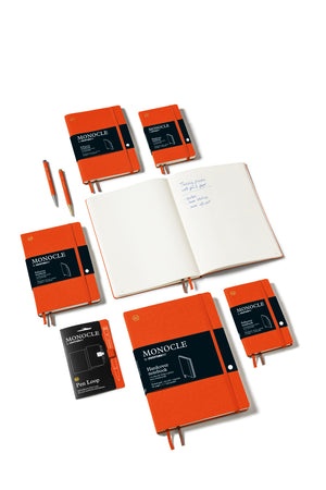 An assortment of orange Monocle notebooks and two orange pens arranged on a clean white background, including closed notebooks with black bands, an open notebook with handwritten text, and a smaller black-and-orange Pen Loop notebook.