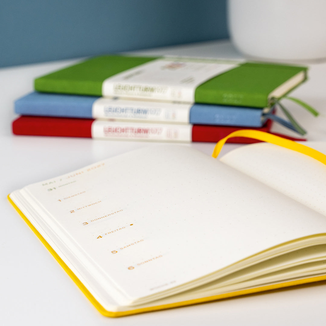 An open daily planner or notebook with blank pages and a yellow ribbon bookmark in the foreground, with three closed notebooks stacked in the background on a white surface.