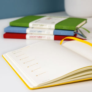 An open daily planner or notebook with blank pages and a yellow ribbon bookmark in the foreground, with three closed notebooks stacked in the background on a white surface.
