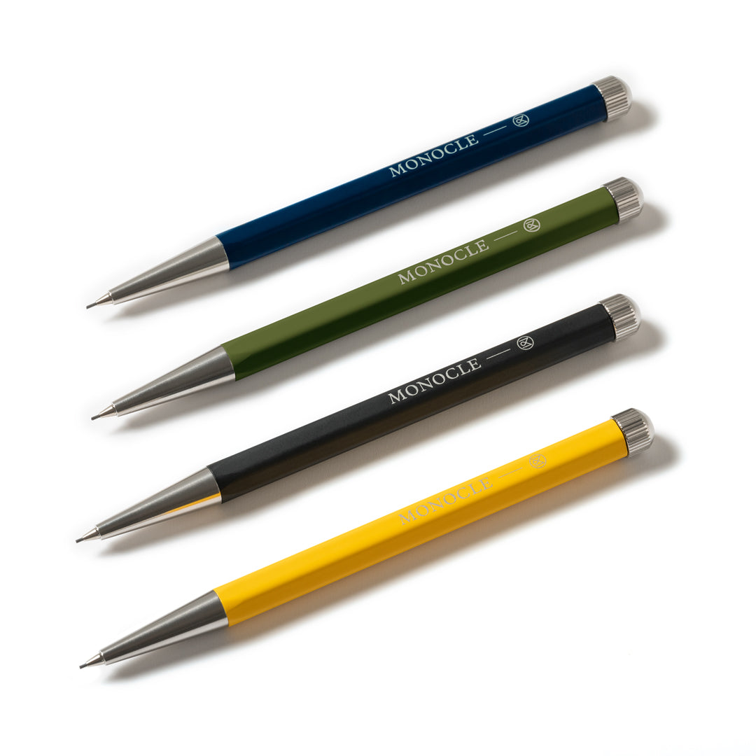 Four Monocle retractable mechanical pencils in navy blue, olive green, black, and yellow, arranged diagonally on a clean white background.