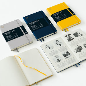 Three closed Monocle notebooks in gray, navy blue, and yellow are arranged on a white surface, alongside two open notebooks showing printed pages and ribbon bookmarks.