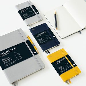 Four Monocle Softcover notebooks in gray, navy, yellow, and white, arranged on a white surface with a black pen and an open notebook.