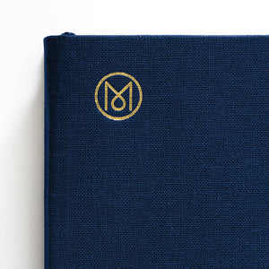 A close-up of a navy blue woven fabric notebook cover with a gold monogram logo on the front, photographed against a clean white background.